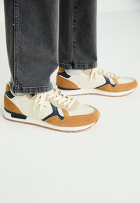 Sneakers feature a combination of cream mesh, tan suede, and navy accents. Rubber sole with contrasting textures; lace-up design.