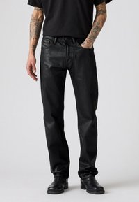 Jeans de mezclilla negra con un corte ajustado, ligero brillo, diseño de cinco bolsillos y herrajes metálicos, combinados con botas de cuero negras.