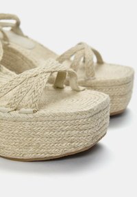 Sandales plateformes en espadrille tissée beiges avec des sangles croisées et une surface texturée. Présentent une épaisse semelle en corde tressée et un fond plat.