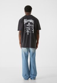 Sort bomulds t-shirt med hvid grafisk design, der viser kors på ryggen; parret med lyseblå brede denim jeans.