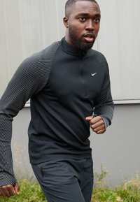 Sort langærmet atletisk top med høj krave, med teksturerede grå striber på ærmerne og et lille hvidt Nike-logo på brystet.
