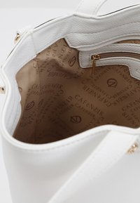Borsa a braccio in pelle bianca con una superficie testurizzata. All'interno presenta una fodera beige con un motivo ripetuto, una tasca con zip e hardware dorato.