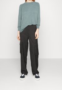 Hellgrüner gestrickter Pullover mit langen Ärmeln, kombiniert mit schwarzen Cargo-Hosen mit Streifen, die seitliche Taschen und weiße Plateausneaker haben.
