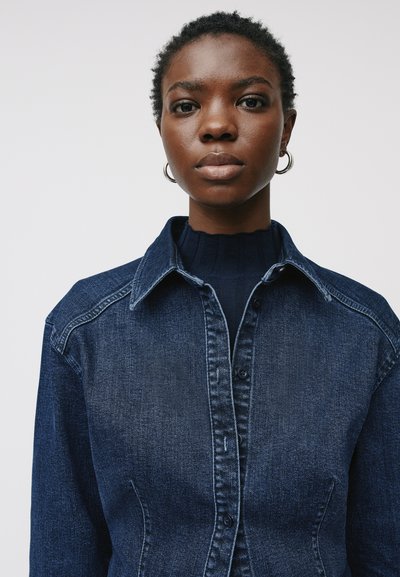 Camicia in denim di colore blu scuro, con chiusura a bottoni frontale, colletto a punta e tessuto resistente. Presenta un design attillato e cuciture in contrasto.