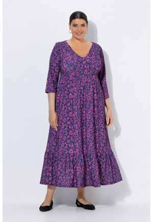 Kvinde med mørkt hår i en lilla og pink blomster V-hals kjole med trekvart ærmer og sorte flade sko, stående mod en hvid baggrund.