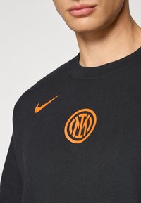 Musta huppari, jossa on pyöreä oranssi logo ja Niken oranssi swoosh rinnan kohdalla. Pyöreä pääntie, valmistettu pehmeästä kankaan koostumuksesta.