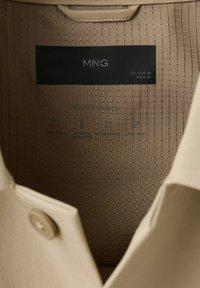 Collet de chemise beige avec un bouton, montrant l'étiquette de la marque MNG et la taille moyenne pour l'UE-USA et le Mexique, avec des icônes d'entretien performance à l'intérieur.