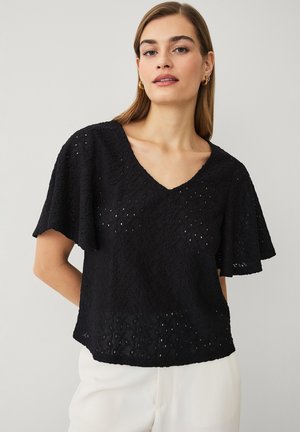 Kobieta ubrana w czarną, teksturowaną bluzkę z krótkim rękawem i dekoltem w serek oraz białe spodnie, stojąca na tle jednolitego, jasnego tła.