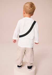 Vit långärmad skjorta med en svart böjd remdetail, beige cargo-byxor och svarta sneakers, sedd bakifrån.