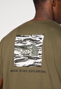Alyvuogių žalios spalvos medvilnės marškinėliai su juodai balta kalnų grafika, kurioje yra "THE NORTH FACE" logotipas ir tekstas "NEVER STOP EXPLORING".