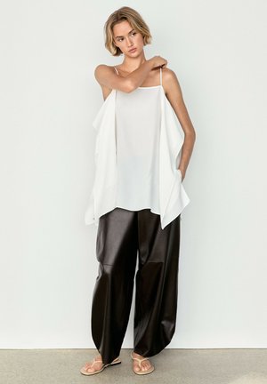 Donna che indossa una canotta bianca larga senza maniche, pantaloni larghi in pelle nera e sandali beige, in piedi davanti a uno sfondo bianco semplice.