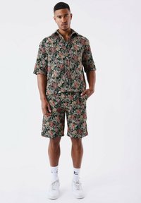 Chemise à manches courtes à motifs floraux et shorts assortis en vert foncé, ornés de roses roses et beiges. Fabriqué en tissu léger, associé à des baskets blanches.