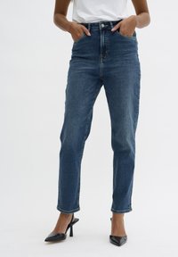 Vaqueros de mezclilla azul oscuro de tiro alto con bolsillos frontales, piernas ajustadas y dobladillos recortados, combinados con tacones negros de punta afilada.