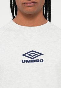 Persona con capelli intrecciati che indossa una maglietta grigio chiaro con il logo Umbro blu navy centrato sul petto.