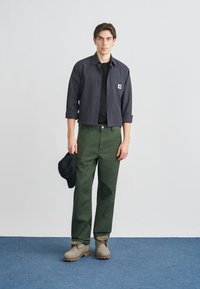 Καθημερινή εμφάνιση με μαύρο καρό πουκάμισο κοντομάνικο, μαύρο μπλουζάκι, πράσινο παντελόνι, γκρι μπότες και μαύρο καπέλο.