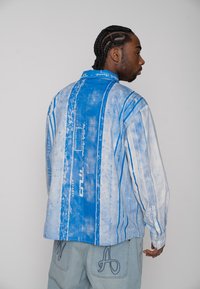 Mężczyzna z warkoczami, ubrany w niebiesko-białą koszulę w paski z długim rękawem oraz jasne dżinsy z dużą haftowaną literą "A" na tylnej kieszeni.