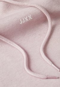 Svijetlo ružičasta tkanina s mekanom teksturom, s pletenim vezicama i utisnutim natpisom "JUXX" u bijeloj boji.