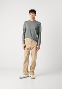 Langärmliges graues Shirt mit kleinem weißen Logo, beige Hose und weiße Sneakers mit beige Akzenten, stehend vor einem weißen Hintergrund.