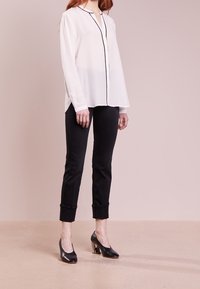 Blouse blanche en chiffon à manches longues avec passepoil noir, associée à un pantalon noir ajusté et des chaussures à talons noires. Texture lisse, coupe moderne.