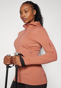 Sweatshirt justificada, com mangas longas e fecho éclair, na cor rosa empoeirada, com um colarinho alto, buracos para os polegares e um bolso lateral. Modelo segurando bastões de caminhada.
