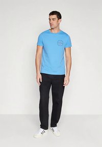 Jasnoniebieska koszulka bawełniana z okrągłym dekoltem i krótkimi rękawami. Na lewej piersi znajduje się okrągły nadruk graficzny. W zestawie z czarnymi spodniami.