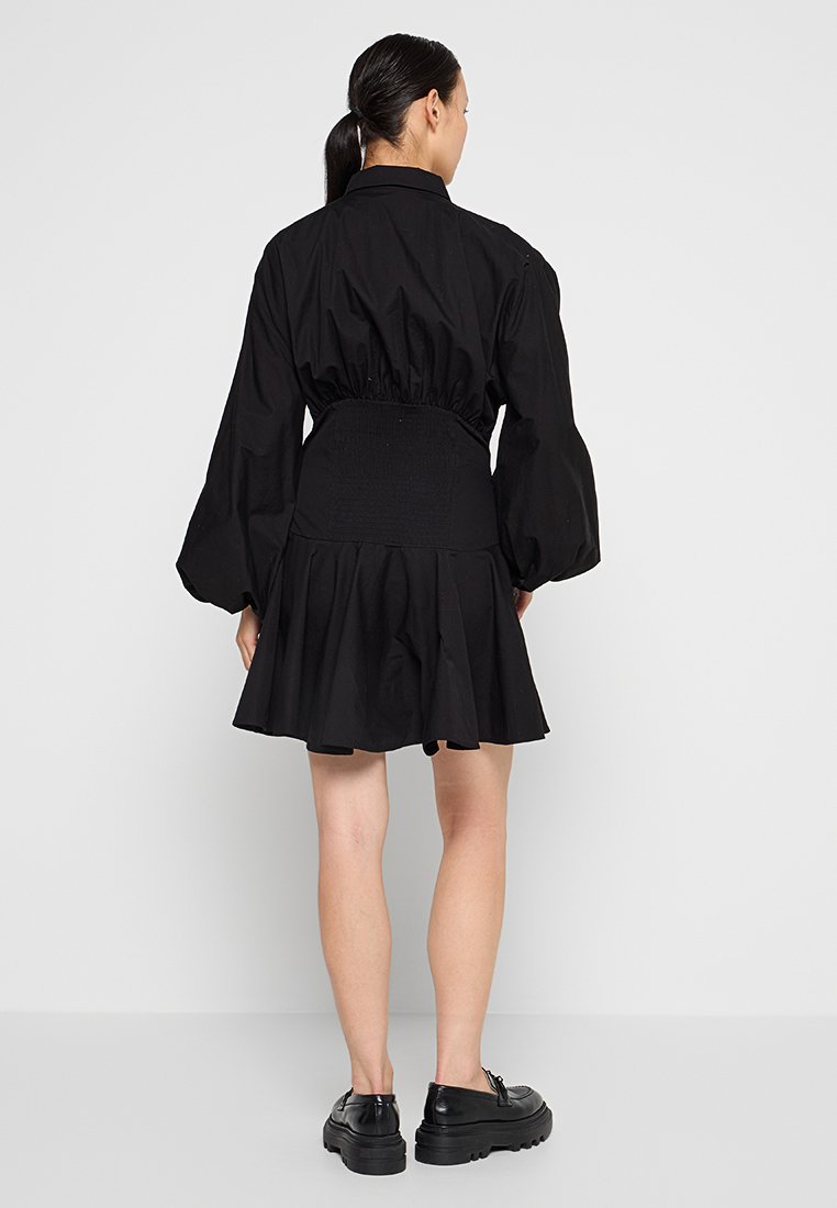 Frau mit schwarzen Haaren in einem Pferdeschwanz, die ein schwarzes langärmliges Kleid mit tailliertem Schnitt und schwarzen klobigen Schuhen trägt, steht vor einem schlichten Hintergrund.