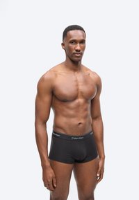 Γυμνασμένος άντρας με κοντά μαλλιά στέκεται με το πρόσωπο προς τα εμπρός, φορώντας μαύρα εσώρουχα Calvin Klein, μπροστά σε απλό ανοιχτό φόντο.