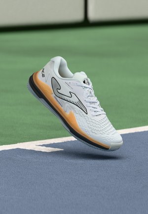 Chaussure de sport blanche avec détail de semelle orange et logo noir flottant au-dessus d'une surface de court de tennis bleu et vert.