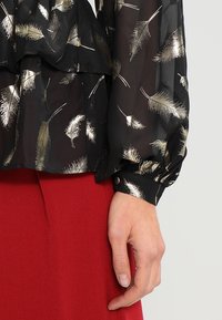 Blusa nera con una texture satinata, caratterizzata da motivi in foglia d'oro metallico. Maniche lunghe con polsini arricciati e dettaglio con bottone.