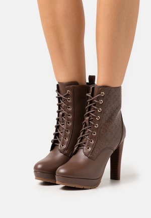 Braune hochhackige Stiefeletten mit vorderen Schnürsenkeln und geprägten Seitenmustern, getragen an nackten Beinen.