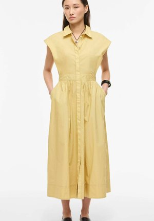 Žena v žltých midi šatách bez rukávov s golierom a zapínaním na gombíky, so vreckami, čiernym náramkom a náhrdelníkom, stojaca na bielom pozadí.