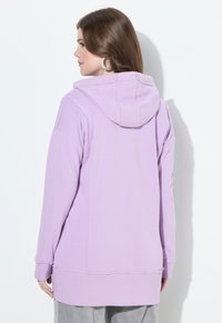 Femme aux cheveux bruns portant de grandes boucles d'oreilles et un sweat à capuche lâche couleur lavande, vue de dos sur fond blanc.