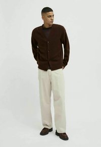 Homem em pé, vestindo um cardigan castanho-escuro abotoado, camisa preta, calças beje claras e sapatos castanhos, contra um fundo simples.