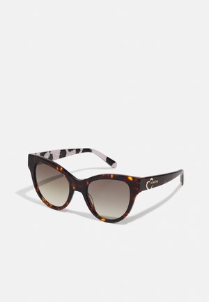 Lunettes de soleil en forme d'œil de chat avec un motif écaille de tortue, des verres dégradés et des branches noires avec des détails blancs et un logo sur le côté.