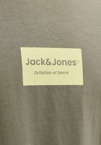 Étiquette en tissu beige clair avec les inscriptions "Jack & Jones" et "Definition of Denim" imprimées sur un fond de tissu gris texturé.