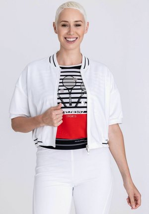 Lächende Person mit kurzen blonden Haaren trägt weiße Jacke über gestreiftem Sportshirt mit Tennisschläger-Motiven und weiße Hose.