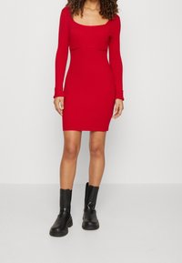 Abito rosso a maniche lunghe con scollatura quadrata, silhouette aderente e orlo al ginocchio. Abbinato a stivali neri alla caviglia.