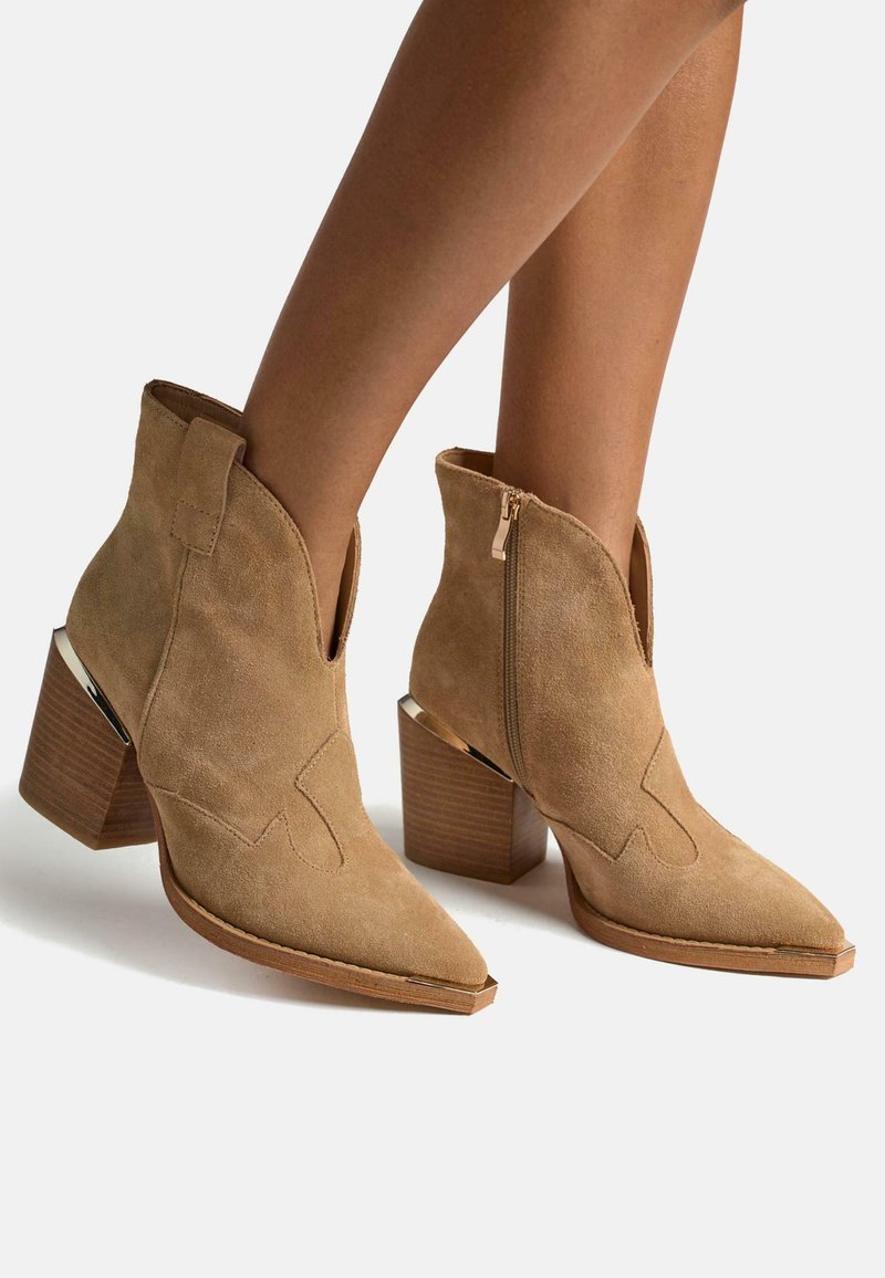 Botines de ante marrón con punta afilada, costuras decorativas, cremallera lateral y tacones de bloque de madera con un acento metálico.
