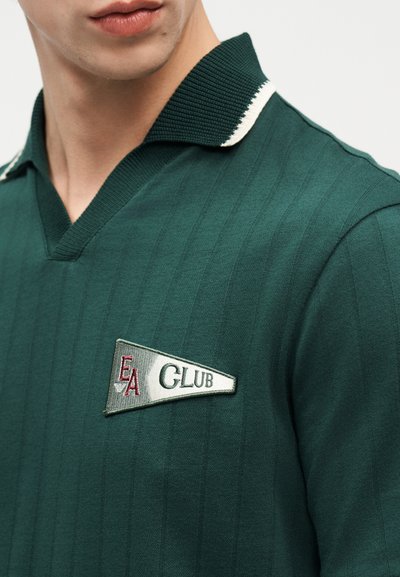 Άνδρας που φοράει πουκάμισο σκούρου πράσινου χρώματος με γιακά λευκής περιμέτρου και μπαλώμα "EA CLUB" στο στήθος.
