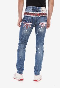 Jean en denim bleu avec un motif éclaboussé, accents de l'Union Jack sur les poches arrière et ceinture à logo. Design ajusté.