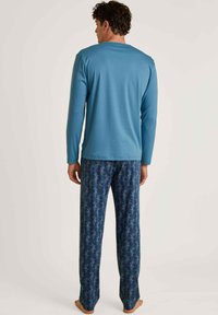 Top blu a maniche lunghe con colletto rotondo e pantaloni da pigiama blu a fantasia con un design foglioso in tonalità più chiare, con piedi nudi visibili.