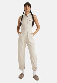 Un mono de pana beige con cuello, cremallera frontal y bolsillos laterales. Combinado con pantuflas grises y calcetines blancos, el modelo lleva una gorra.