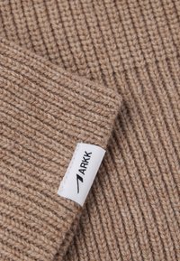 Acercamiento de tela de punto acanalado beige con una etiqueta blanca cosida en la costura que dice "ARKK".