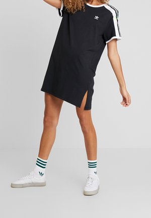 Personne portant une robe T-shirt noire Adidas avec des bandes blanches et un logo, assortie à des chaussettes blanches Adidas et des baskets blanches.