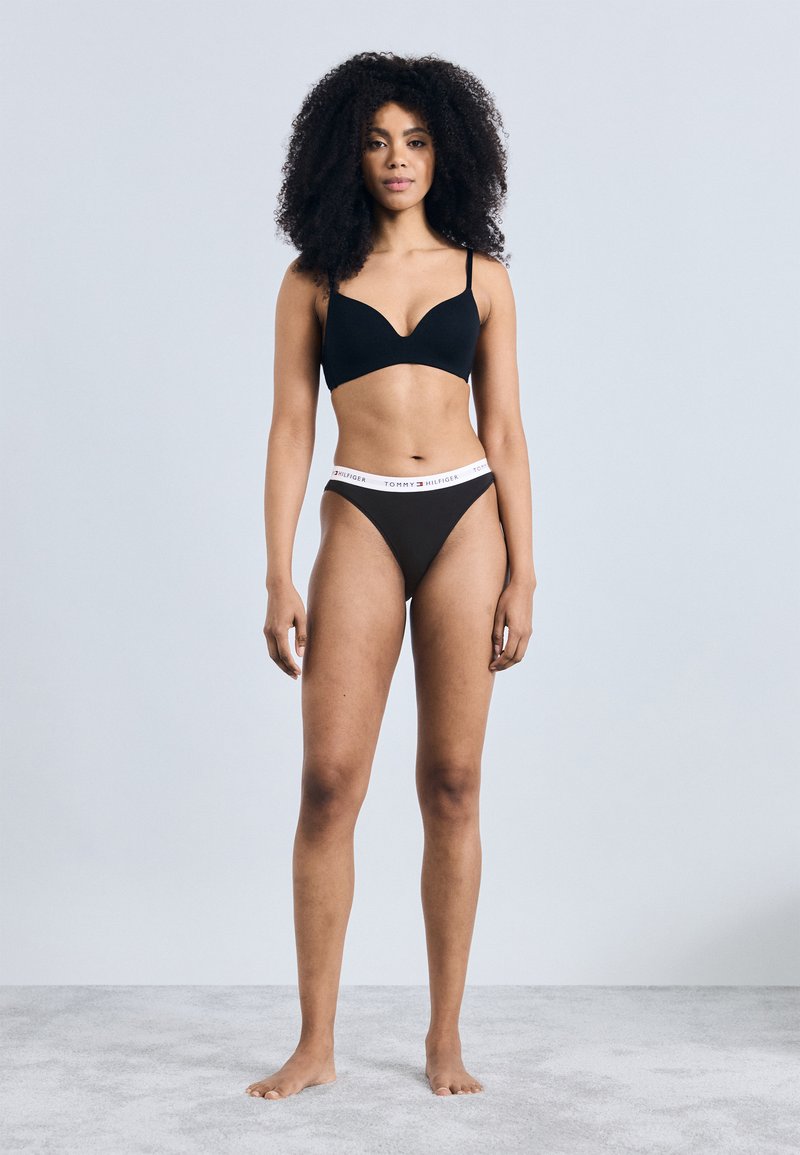 Μαύρο ασύρματο σουτιέν με λεία επιφάνεια, συνδυασμένο με μαύρο στρινγκ που έχει λευκή μέση με λογότυπο. Το μοντέλο στέκεται ξυπόλητο σε ανοιχτόχρωμη επιφάνεια.