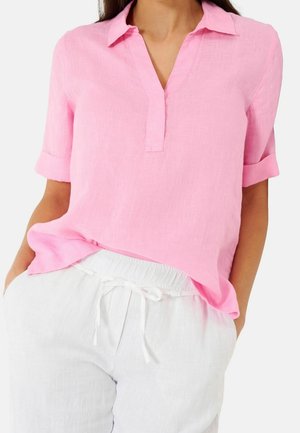 Mujer con una camisa de lino rosa claro con botones hasta la mitad y mangas remangadas, y pantalones de lino blancos con cordón y las manos en los bolsillos.