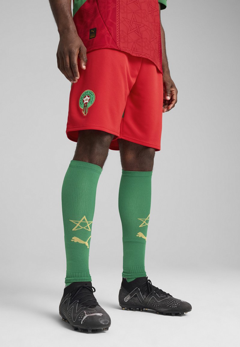 Röd fotbollsjersey med gröna detaljer, röda shorts, gröna strumpor med mönster av gula stjärnor och svarta fotbollsskor med silverdetaljer.