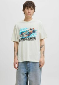 Tricou bej deschis din bumbac, cu un print grafic al unei mașini de curse și text. Are mâneci scurte și o croială lejeră, asociat cu blugi albaștri deschiși.