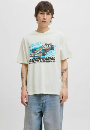 Lys beige T-shirt i bomuld med et grafisk print af en racerbane og tekst. Har korte ærmer og en løs pasform, parret med lyseblå jeans.