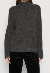 Pull en tricot à côtes gris avec un col montant et des manches longues, fabriqué à partir d'un matériau texturé. Assorti à un pantalon en velours côtelé noir.